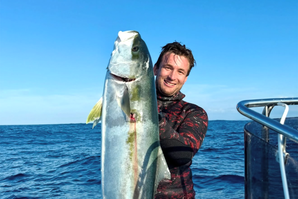 Person on a boat holding up a large freshly caught fish, smiling with the ocean in the background.