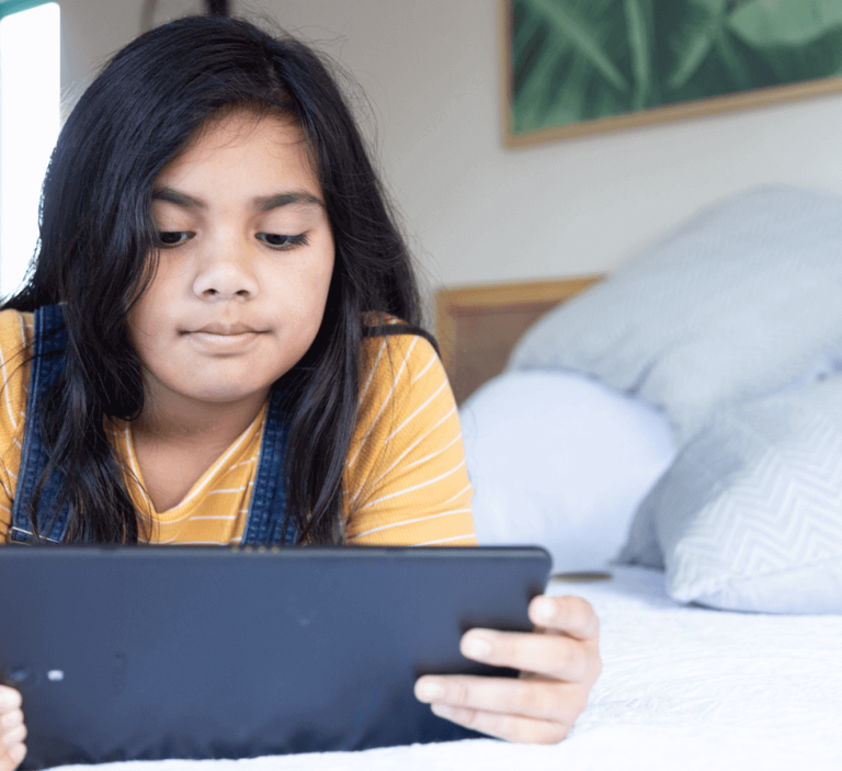 Girl lies on bed looking at a digital tablet.