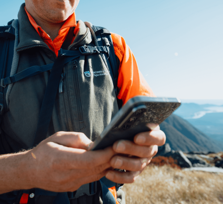 A person wearing an orange shirt and gray vest with a backpack is using a smartphone.