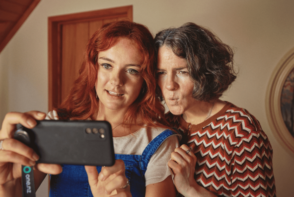 Two people taking a selfie indoors with a smartphone, smiling at the screen in a warm, casual home setting.