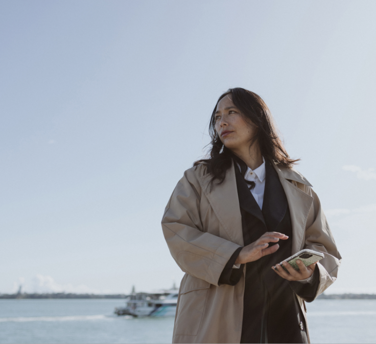 A business woman holding a phone looking to the distance with a harbour behind her.